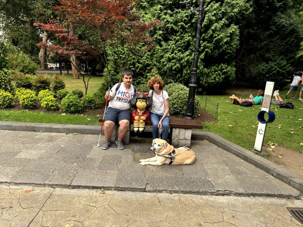 Dos personas sentadas en un banco de parque junto a una escultura de Mafalda. El hombre a la izquierda tiene barba corta, viste una camiseta clara con la palabra "HIGH" en letras grandes, pantalón corto y sostiene un bastón blanco y rojo. La mujer a la derecha lleva camiseta blanca, pantalones y sujeta la correa de un perro guía, que está acostado en el suelo frente a ella. Detrás, hay árboles, arbustos y otras personas tumbadas sobre el césped. Parece un ambiente relajado en un parque urbano.