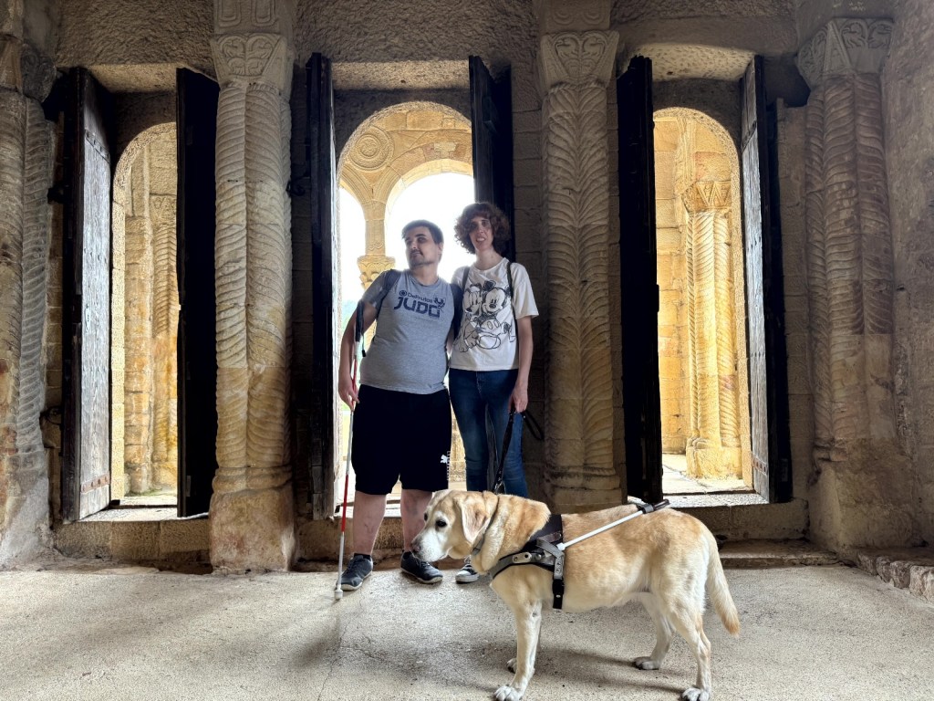 Dos personas de pie una junto a la otra, acompañadas por un perro guía que está delante de ellas. Están en el interior de un edificio antiguo de piedra, decorado con columnas talladas y arcos. Detrás hay varias puertas o ventanas abiertas por donde entra la luz natural, iluminando el espacio y enfatizando las texturas de la arquitectura histórica. El ambiente transmite una sensación de tranquilidad y antigüedad, y el perro parece atento y en reposo.